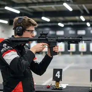 Person at indoor shooting range aiming rifle in shooting stance, wearing protective gear.
